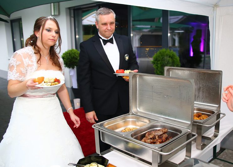 Hochzeitsfeier mit Braut und Bräutigam vor einem Buffet mit warmen Speisen.