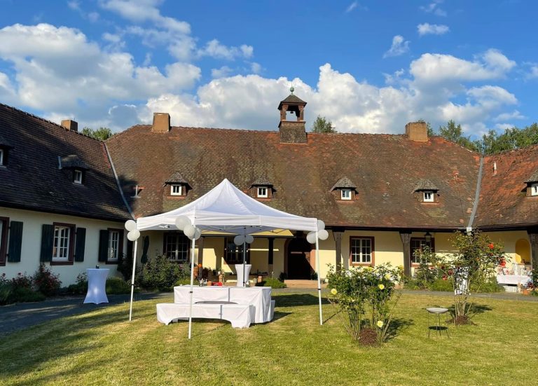 Weißes Zelt und Dekoration in einem Garten vor einem traditionellen Gebäude unter blauem Himmel.